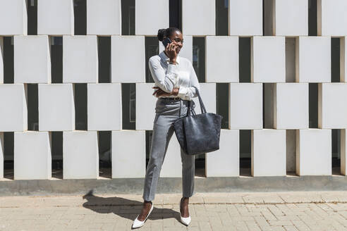 Mid adult businesswoman with purse talking on mobile phone during sunny day - JRVF00579