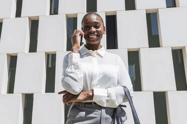 Smiling professional talking on mobile phone while standing in front of block shape wall - JRVF00581