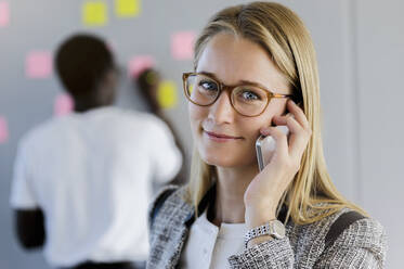 Young female entrepreneur talking on mobile phone with male colleague working in background at office - BMOF00595