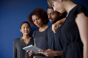 Geschäftsfrau mit Blick auf internationale männliche und weibliche Kollegen, die an einem digitalen Tablet vor blauem Hintergrund arbeiten - BMOF00606
