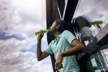 Young man drinking water while leaning on glass wall - BMOF00611