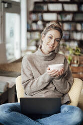 Female owner holding bowl while sitting with laptop on bowl at cafe - JOSEF04401