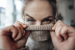 Playful woman covering face in sweater at cafe - JOSEF04412