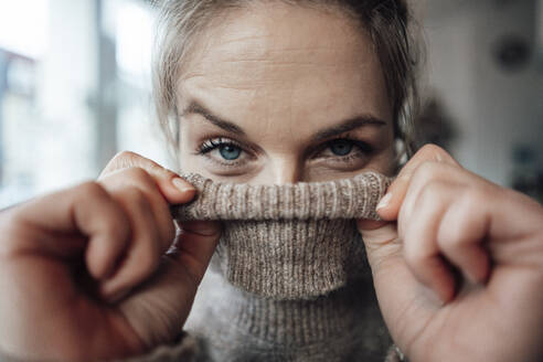 Playful woman covering face in sweater at cafe - JOSEF04412