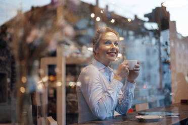 Thoughtful female entrepreneur with coffee cup smiling while looking through cafe window - JOSEF04423