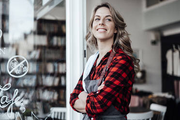 Confident female entrepreneur with arms crossed standing at cafe door - JOSEF04457