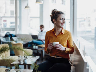 Lächelnde Geschäftsfrau hält eine Kaffeetasse, während sie mit einem Kollegen in einem Café sitzt - JOSEF04513