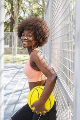 Lächelnde Frau mit lockigem Haar und Basketball, der an einem Zaun lehnt - IFRF00571