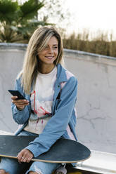 Lächelnde Frau mit Skateboard, die wegschaut, während sie ein Mobiltelefon im Park hält - XLGF01791