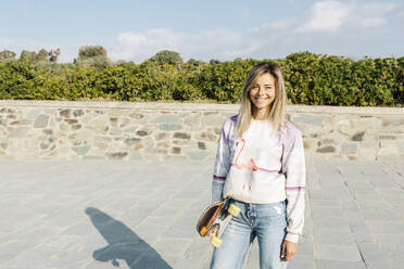 Smiling woman with skateboard standing in park during sunny day - XLGF01804