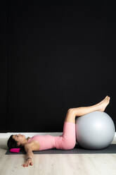 Sportlerin auf Übungsmatte mit Fitnessball im Studio liegend - GIOF12637