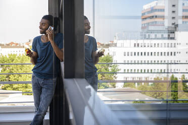 Lächelnder Mann, der mit seinem Smartphone spricht, während er auf dem Balkon einen Apfel isst - BMOF00658