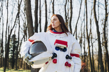 Nachdenkliche Astronautin mit Weltraumhelm, die im Wald in die Ferne schaut - MEUF02814