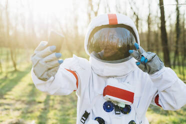 Weibliche Astronautin in Raumanzug und Helm, die ein Selfie mit ihrem Mobiltelefon im Wald macht - MEUF02831