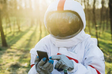 Junger Astronaut mit Raumanzug und Helm, der im Wald ein Mobiltelefon benutzt - MEUF02832