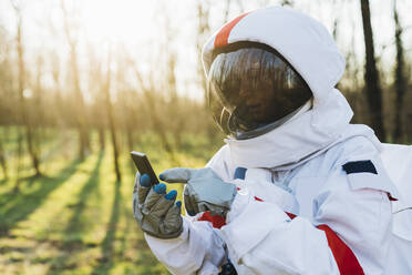 Junger Astronaut mit Raumanzug und Helm benutzt sein Smartphone im Wald - MEUF02833