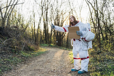 Weibliche Astronautin im Raumanzug gestikuliert, während sie einen Karton auf einem Feldweg hält - MEUF02844