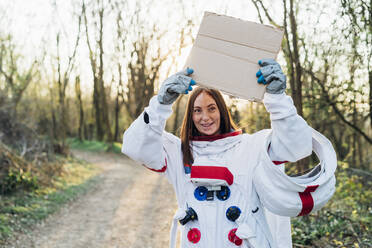 Lächelnder junger Astronaut, der einen Karton in der Hand hält, während er auf einem Feldweg steht - MEUF02845