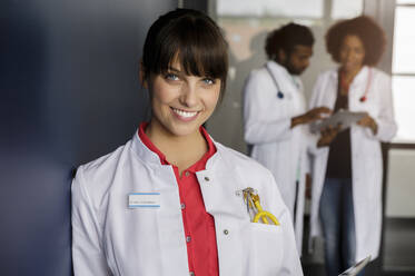 Lächelnde Ärztin mit männlichen und weiblichen Kollegen im Hintergrund im Krankenhaus - BMOF00703
