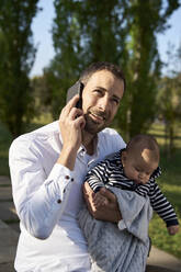 Lächelnder Vater mit einem kleinen Jungen, der wegschaut, während er im Park mit dem Handy telefoniert - VEGF04438