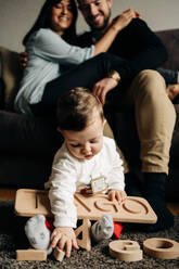 Unbekannte junge Eltern umarmen sich auf dem Sofa in der Nähe des süßen kleinen Sohnes, der auf dem Boden mit Holzspielzeug Buchstaben spielt - ADSF24196