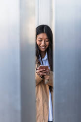 Voller Länge der glücklichen asiatischen Frau in stilvollem Outfit stehen auf der Straße und Surfen im Internet auf dem Handy - ADSF24204