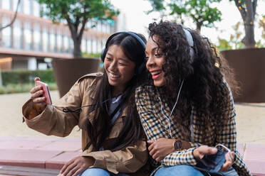 Asian female having fun call on smartphone and focused black woman listening to music and surfing internet on mobile phone - ADSF24206