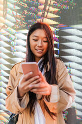 Glückliche asiatische Frau in trendigem Outfit, die ihr Handy an einer digitalen Wand betrachtet - ADSF24209