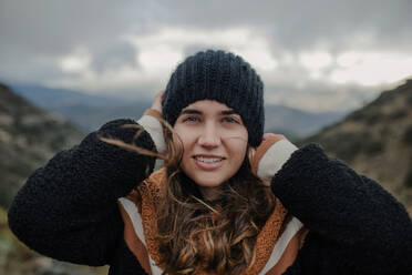 Lächelnde junge Frau, die sich eine warme Mütze aufsetzt und in die Kamera schaut, während sie auf dem rauen Hochland an einem bewölkten, düsteren Tag steht - ADSF24212