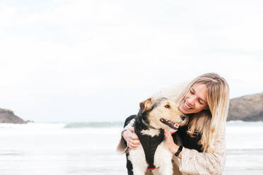 Seitenansicht einer Frau, die einen niedlichen Rassehund umarmt, während sie sich gegenseitig gegen das Meer unter bewölktem Himmel anschaut - ADSF24240