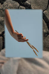 Anonymous female holding grass spikes in hand reflected in mirror against blue sky in nature with stony background - ADSF24243