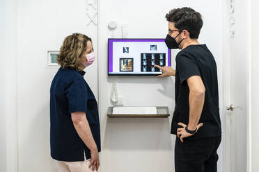 Männlicher Zahnarzt im Gespräch mit einer Frau am Computerbildschirm in einer Klinik während einer Pandemie - DLTSF01887