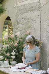 Lächelnde reife Frau, die bei einer Blumenpflanze steht und Brot im Hinterhof zubereitet - ALBF01611