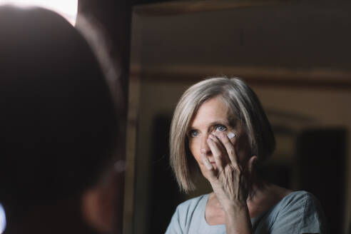 Short haired woman looking in mirror while applying face cream at home - ALBF01632