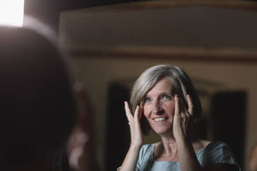 Lächelnde Frau mit Kopf in den Händen, die zu Hause in den Spiegel schaut - ALBF01635