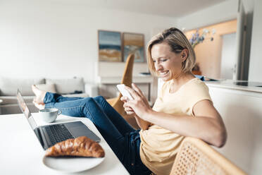 Smiling businesswoman with feet up using mobile phone while working at home - JOSEF04528