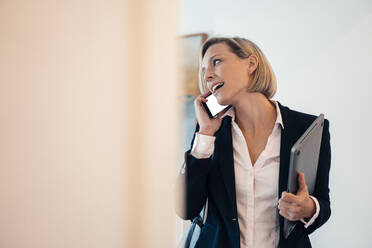 Smiling businesswoman holding laptop while talking on phone at home - JOSEF04542