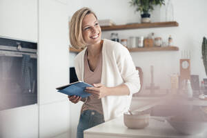 Smiling woman looking away while leaning on kitchen counter at home - JOSEF04553