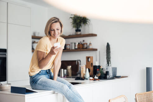 Beautiful smiling woman sitting on kitchen counter at home - JOSEF04554
