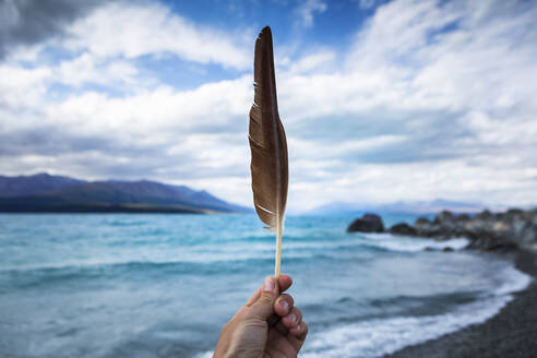 Neuseeland, Canterbury, Hand hält Feder mit Lake Tekapo im Hintergrund - WVF01898