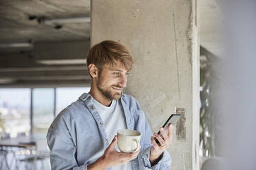 Blond man with coffee cup using mobile phone leaning on column at loft - FMKF07124