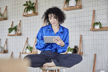 Curly haired woman sitting on stool while using digital tablet - FMKF07128