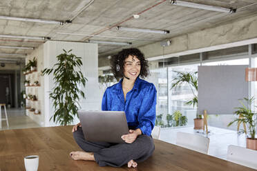 Lächelnde Frau, die wegschaut, während sie mit ihrem Laptop auf dem Tisch im Loft sitzt - FMKF07136