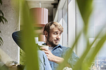 Handsome man with hand in hair looking away while sitting at loft - FMKF07143