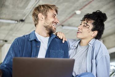 Smiling man with laptop looking at girlfriend in loft - FMKF07148