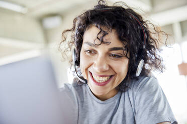 Fröhliche Frau mit Kopfhörern, die auf einen Laptop schaut - FMKF07170