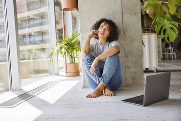 Young woman looking away while sitting on floor at home apartment - FMKF07174
