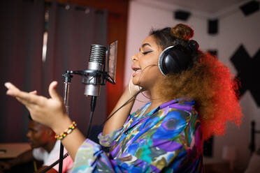 Side view of young African American female vocalist in headphones recording song against smiling music producer in studio - ADSF24258