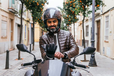 Älterer bärtiger hispanischer männlicher Motorradfahrer mit Schutzhelm, der Handschuhe auf einem zeitgenössischen Motorrad auf der Straße anzieht - ADSF24262