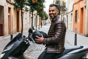 Seitenansicht eines zufriedenen männlichen hispanischen Motorradfahrers mit Helm, der auf einem Motorrad in der Stadt wegschaut - ADSF24264